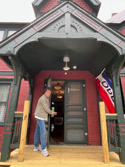 Woman on a small porch opening the door to a historic building with an open flag hanging outside.