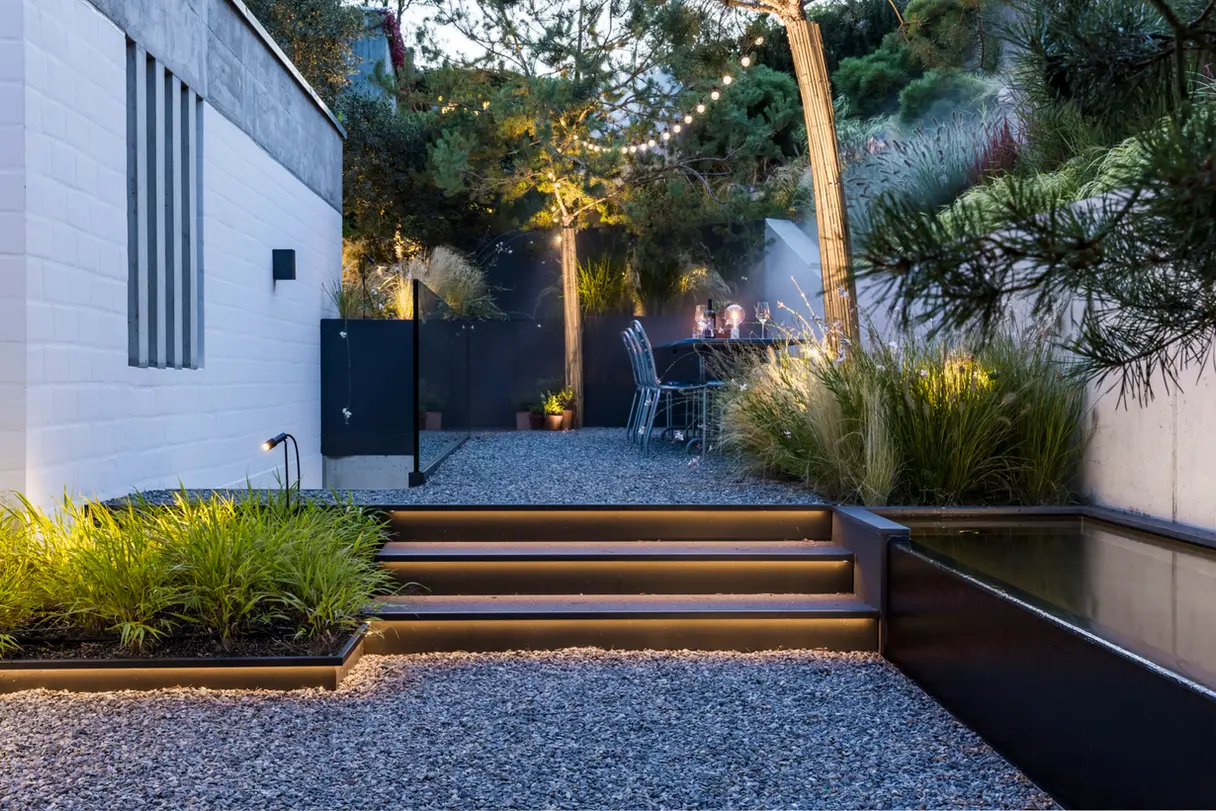 Hanggarten in Bern mit einer breiten Treppe, beleuchtet mit warm leuchtenden LED-Streifen, die zum Essbereich führt. Ein Pool im Vordergrund, Pinien, Stauden und Gräser im Hintergrund.