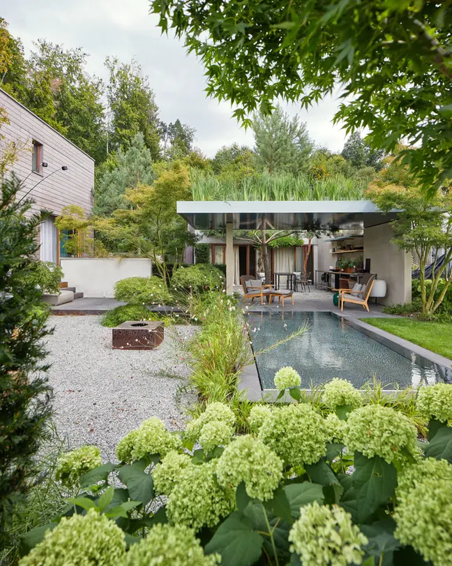 Gartengestaltung in Bern mit einer begrünten Pergola, einem Mosaik-Pool und Feuergrill, Hortensien im Vordergrund und Wald im Hintergrund.