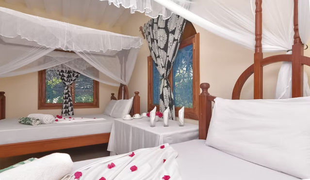 Twin canopy beds with mosquito nets and traditional wood carvings, facing a small white table with flowers.