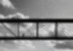 Silhouette d’un piéton marchant sur une passerelle métallique, sous un ciel nuageux en noir et blanc.