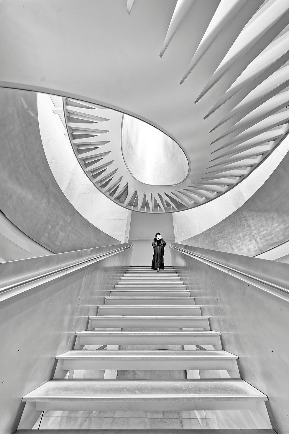 Escalier moderne en noir et blanc, personne en long manteau au sommet, architecture courbe et lumière apaisante.