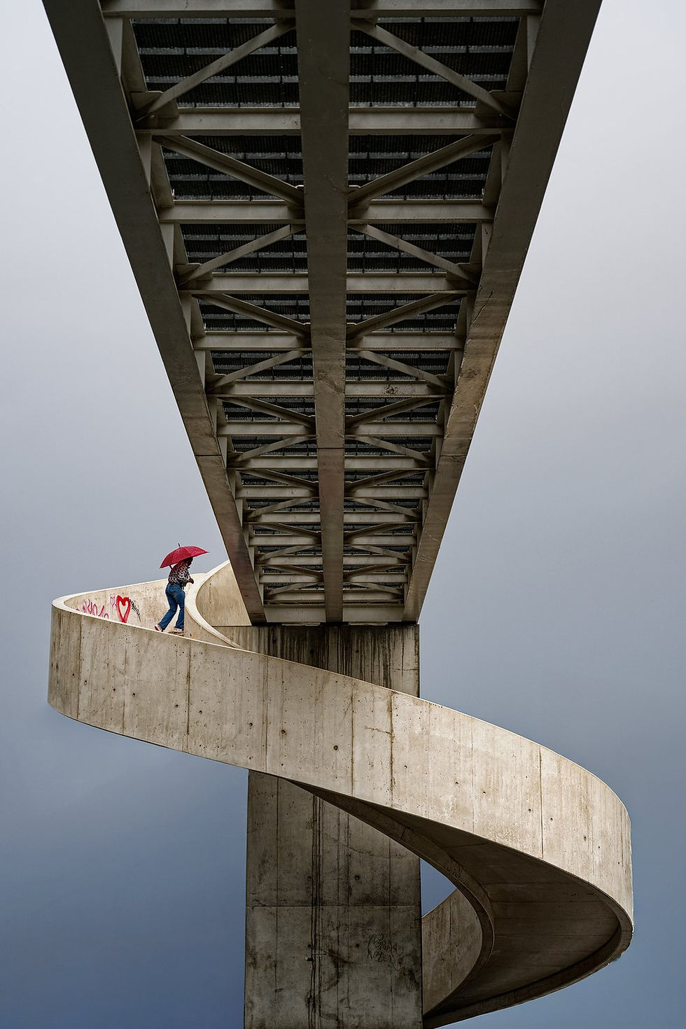 Sous un pont, une personne au parapluie rouge monte un escalier en spirale de béton, avec un cœur graffiti, ciel gris.