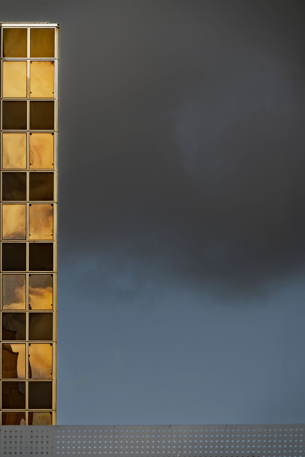 Façade de bâtiment aux fenêtres dorées, à gauche, sous un ciel gris sombre; ambiance calme et austère.