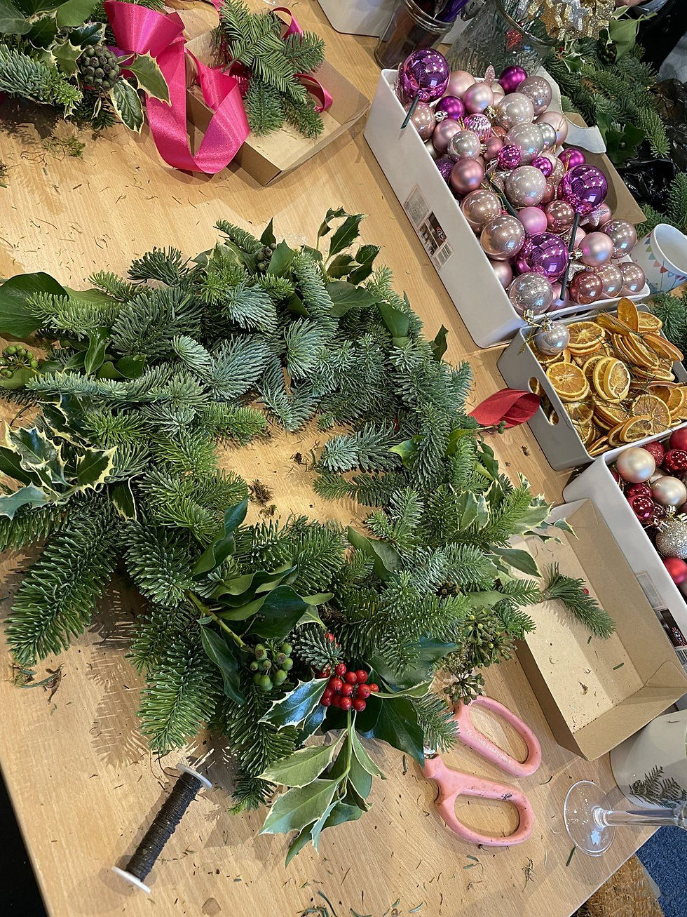 Bundles of fresh fir and holly laid out around a moss ring on a workshop table