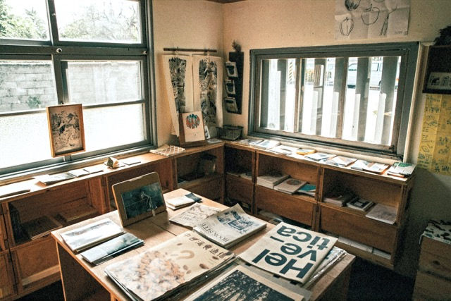 Sunlit printmaking art studio room with wooden shelves, books, and artistic prints. Posters and drawings on the wall. Cozy and creative atmosphere.