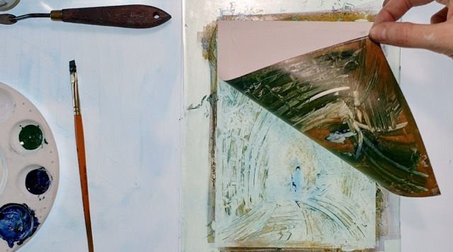 Statira's hand lifts a brown and green toned print from a flat surface reflecting planographic printmaking. Nearby are a paint palette with green and blue, a brush, and a palette knife on a light surface. The flat surface is a gelliplate and the print is a gelliprint.