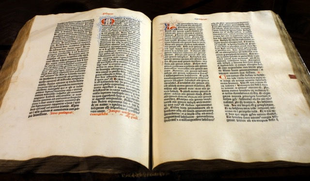 Open ancient Gutenberg bible book with dense black text on yellowed pages, red and blue decorative initials, lying on a wooden table. The page is showcasing intricate blackletter type and detailed illuminated initials, exemplifying the artistry and craftsmanship of early printed books.