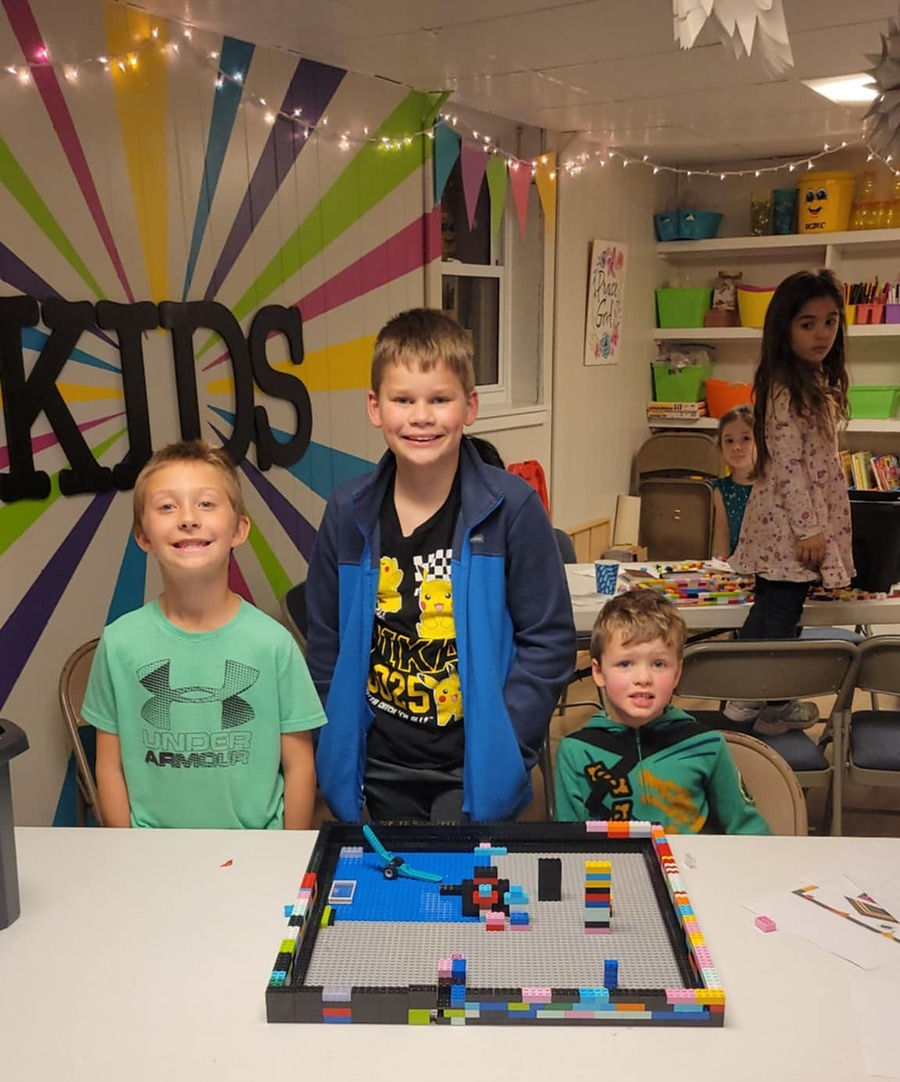 Three boys smiling with a Lego creation in a room marked 'KIDS'