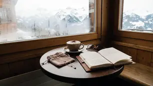 Café-Tisch am Fenster mit Notizbuch, Cappuccino und zwei Layering-Ketten im Beutel, Avirelle.