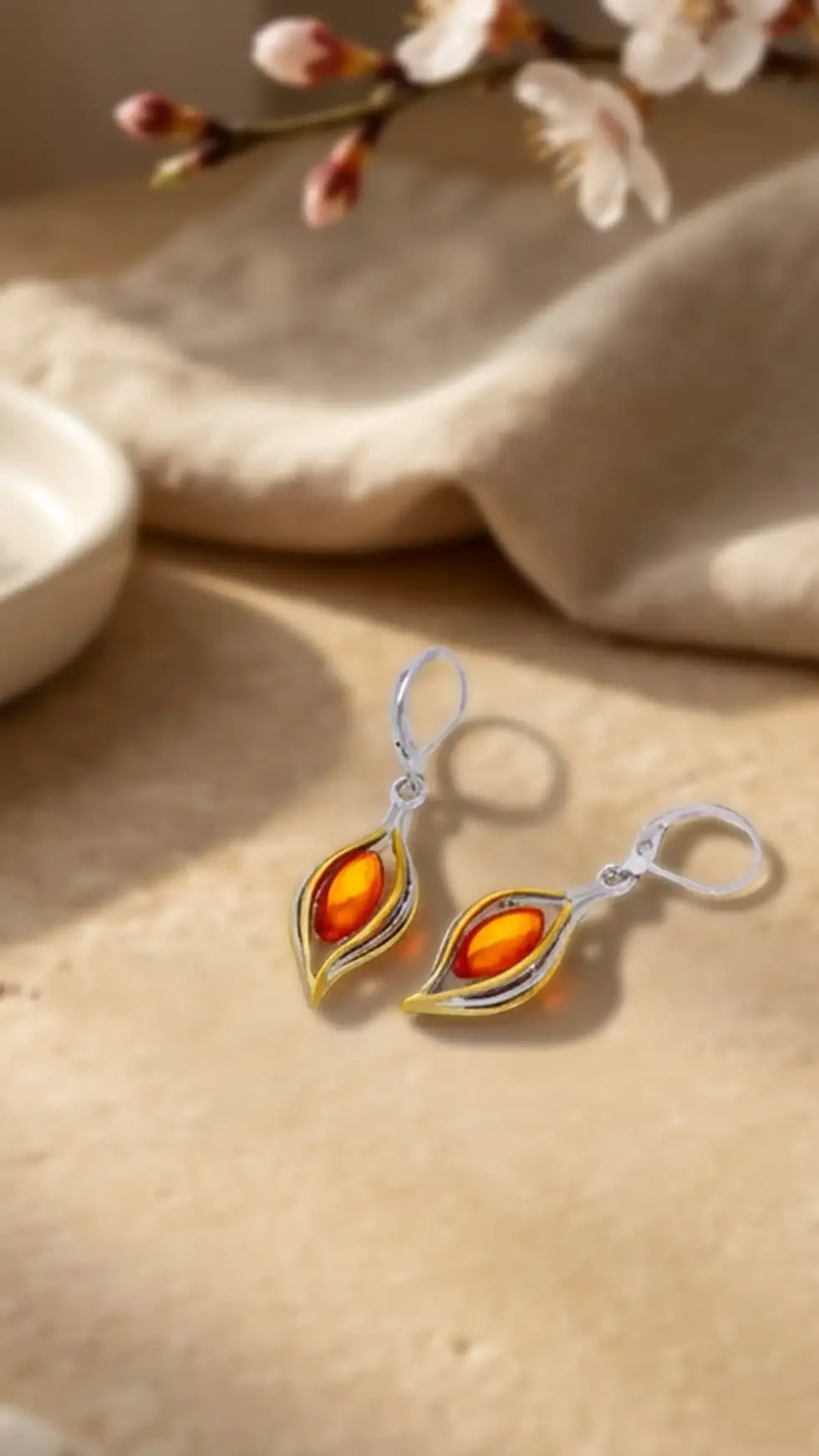 Ohrhänger mit orangerotem Stein im Frühlingslicht auf heller Steinfläche, mit zarten Blüten im Hintergrund, Avirelle.