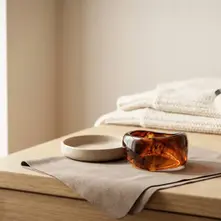 Breiter Acryl-Armreif in brauner Optik auf Mikrofasertuch neben Wollschal, Parfum und Handcreme – Winterpflege. Avirelle.