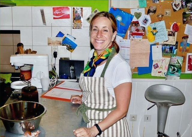 Smiling woman in a kitchen with white tiles, wearing an apron and colorful scarf. Bulletin board with notes behind. Mixing bowl on counter.