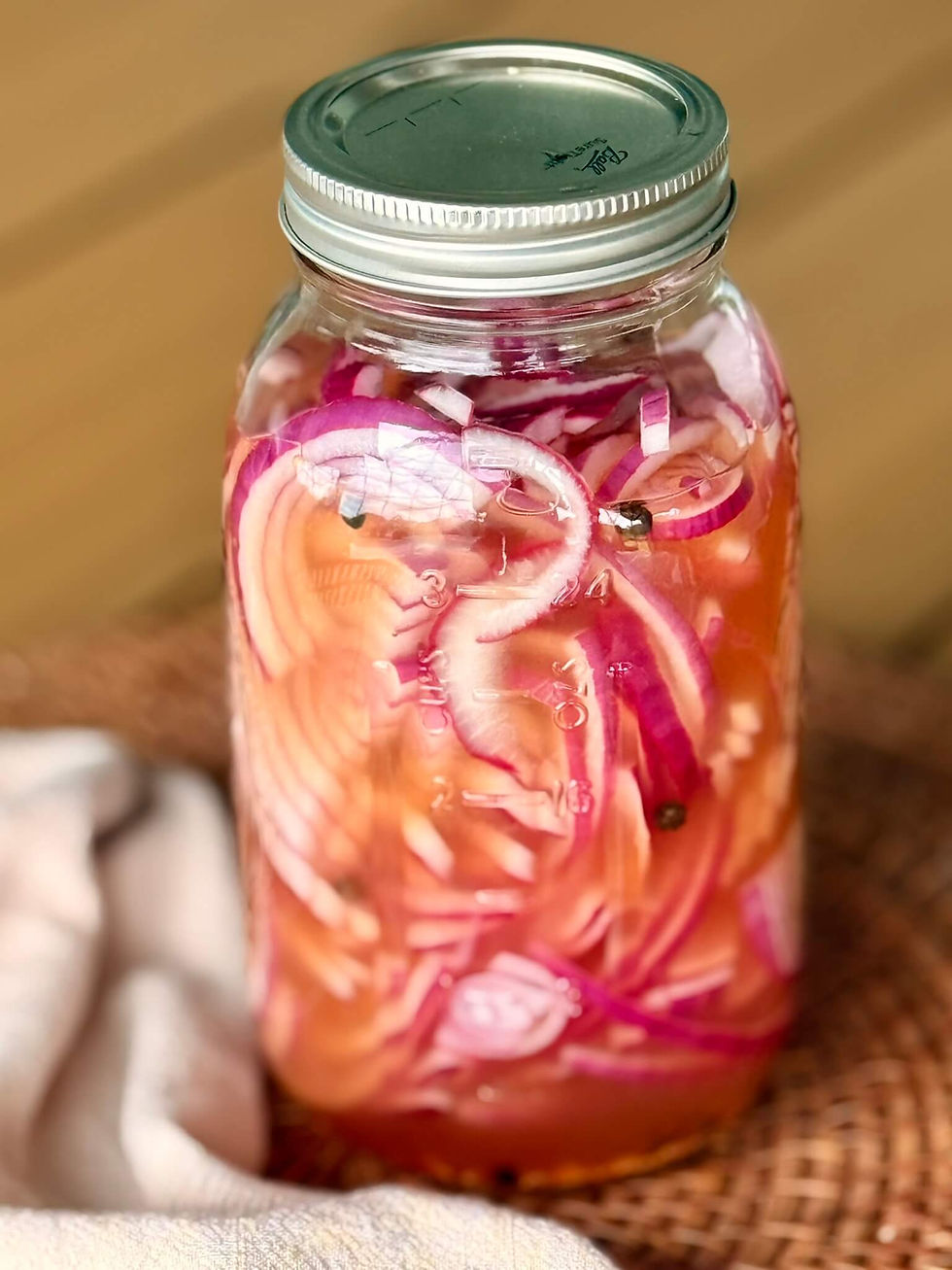 Jar of pickled red onions with liquid, on a woven mat. Metal lid on jar. Soft, neutral-colored cloth nearby. Bright, fresh look.