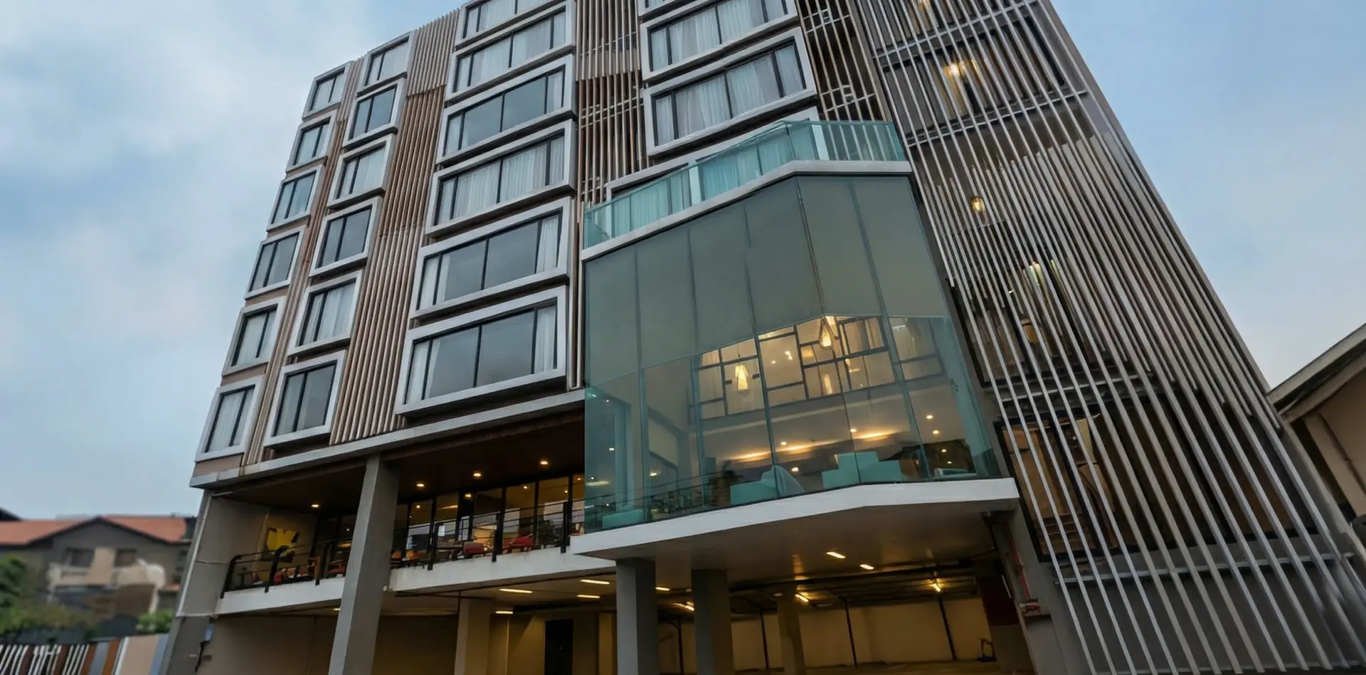 Main street façade of B2 Bangna Premier Hotel showing modular guestroom grid, vertical aluminium fins, and projecting glazed public lobby volume in Bangkok.