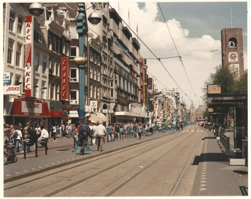 Busy street scene in a European city. People walking along storefronts, including KFC. Tram tracks run down the center. Clock tower in background.