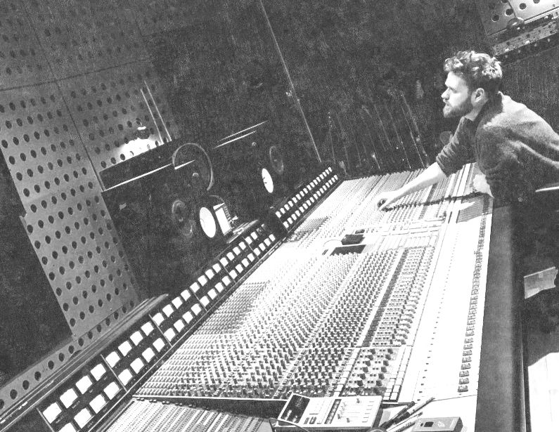 Man in a black-and-white recording studio leans over a huge mixing console, adjusting controls beside speakers.