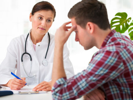A female doctor speaks to a male patient sitting in a chair with his head in his hand looking frustrated.