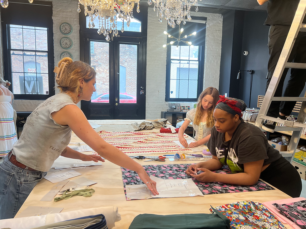 Notice Co-Teacher and Youth Lead Iz Eagles guiding students as they lay out patterns on an unfinished cutting table, meanwhile a parent/volunteer works on a ladder in the background...