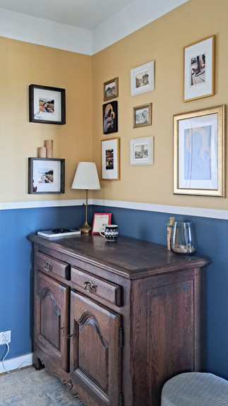 drewniana komoda w przedpokoju, nad którą znajduje się kolaż zdjęć, oraz świętych obrazków na tle żółto-niebieskiej ściany.