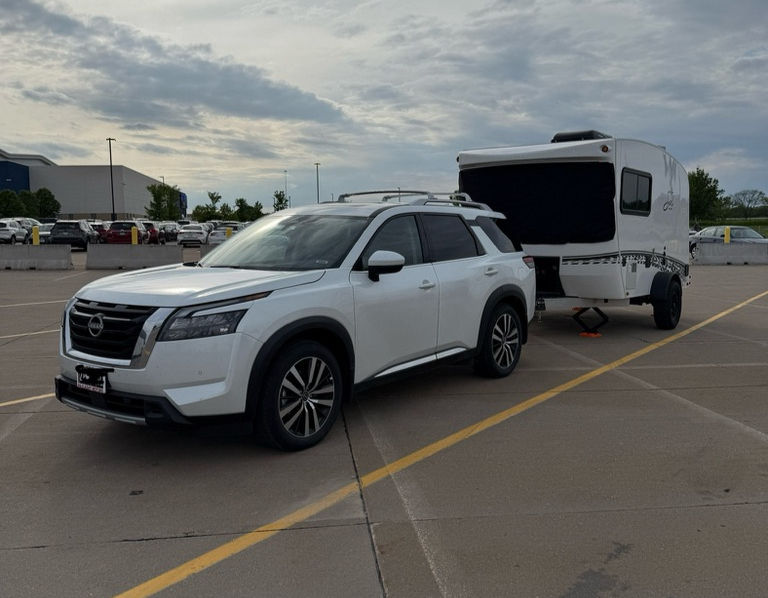 White SUV towing a small trailer parked in an empty lot under a cloudy sky. Adjacent buildings and rows of parked cars in the background.