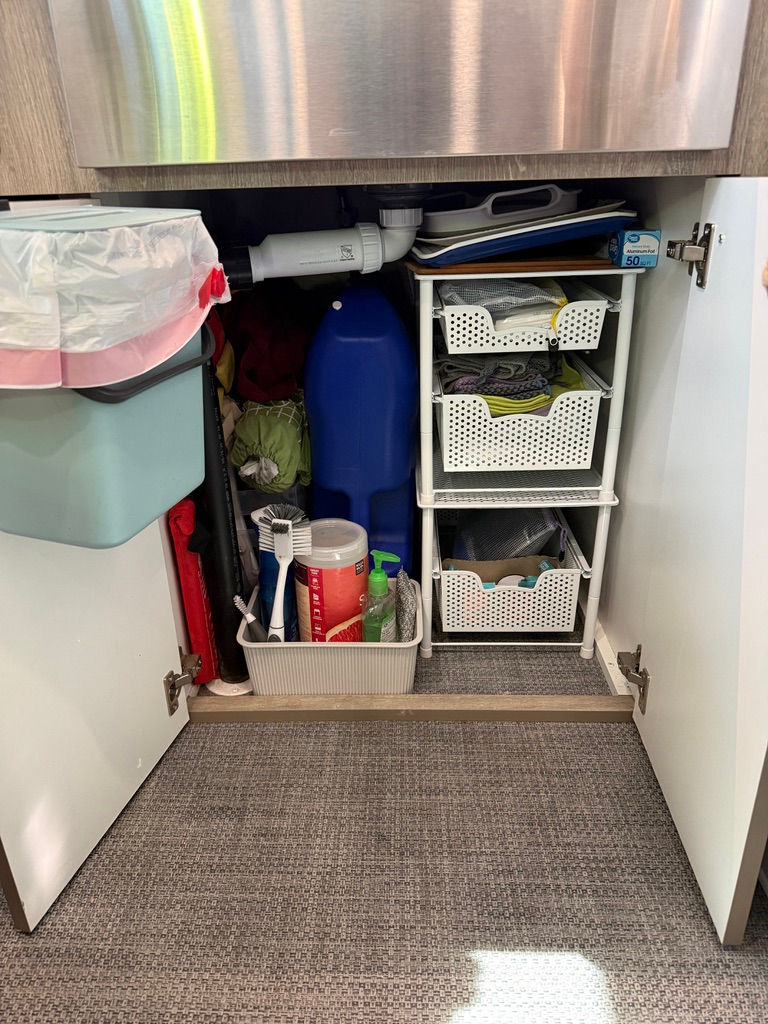 Under-sink cabinet with open doors. Inside are cleaning items, a blue bottle, organized bins, and pipes. A trash bin is attached to the door.