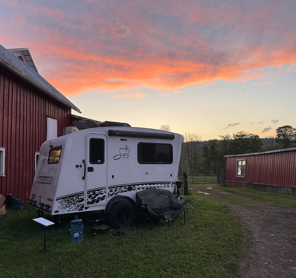 A white camper by a red barn at sunset. The sky is pink and orange. Camping chairs and a water jug are on the grass. Peaceful setting.