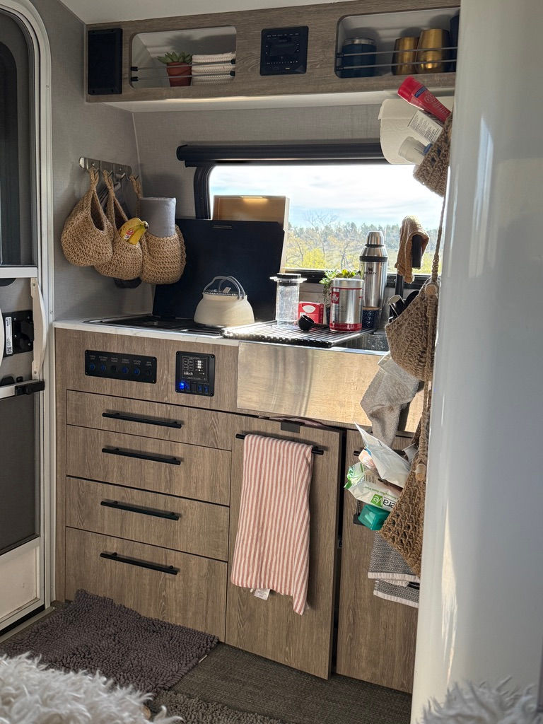 Cozy camper kitchen with wood cabinets, striped towel, and countertop items. Window view of trees and sky. Neutral tones and woven baskets.
