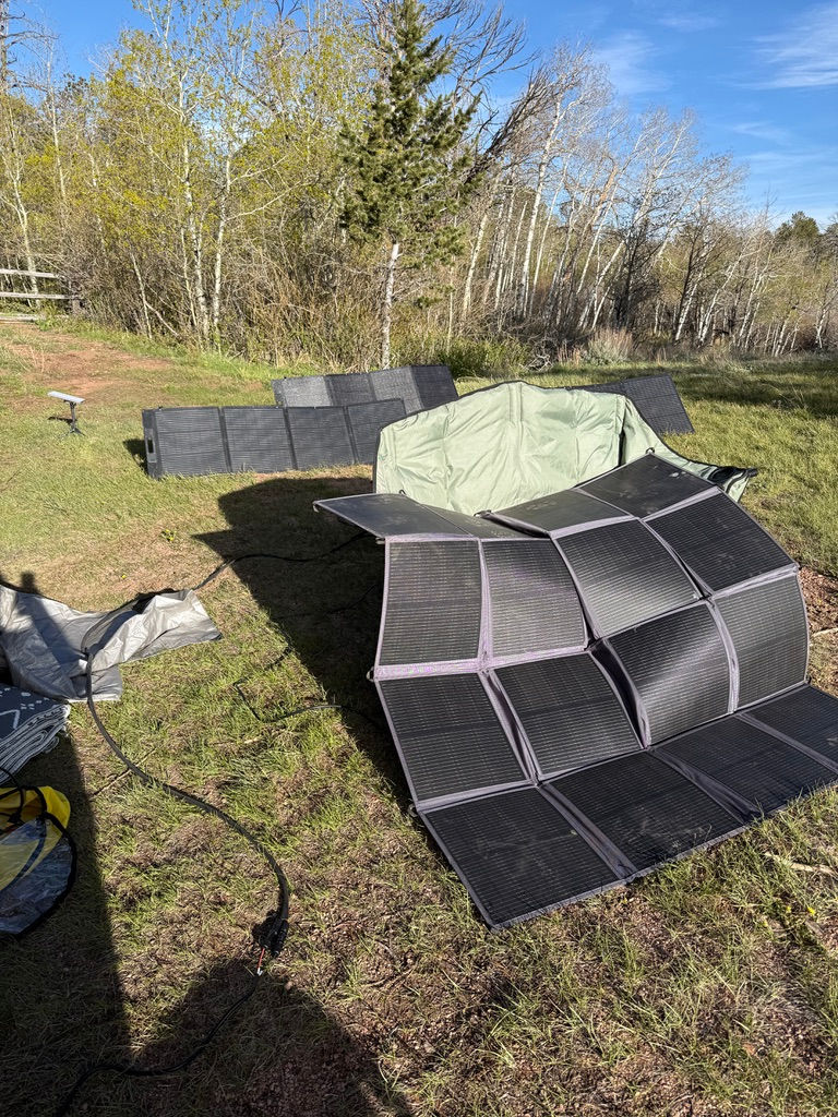 Solar panels unfold on grassy field with trees in background. Green tarp covers some panels. Clear blue sky, sunny day.