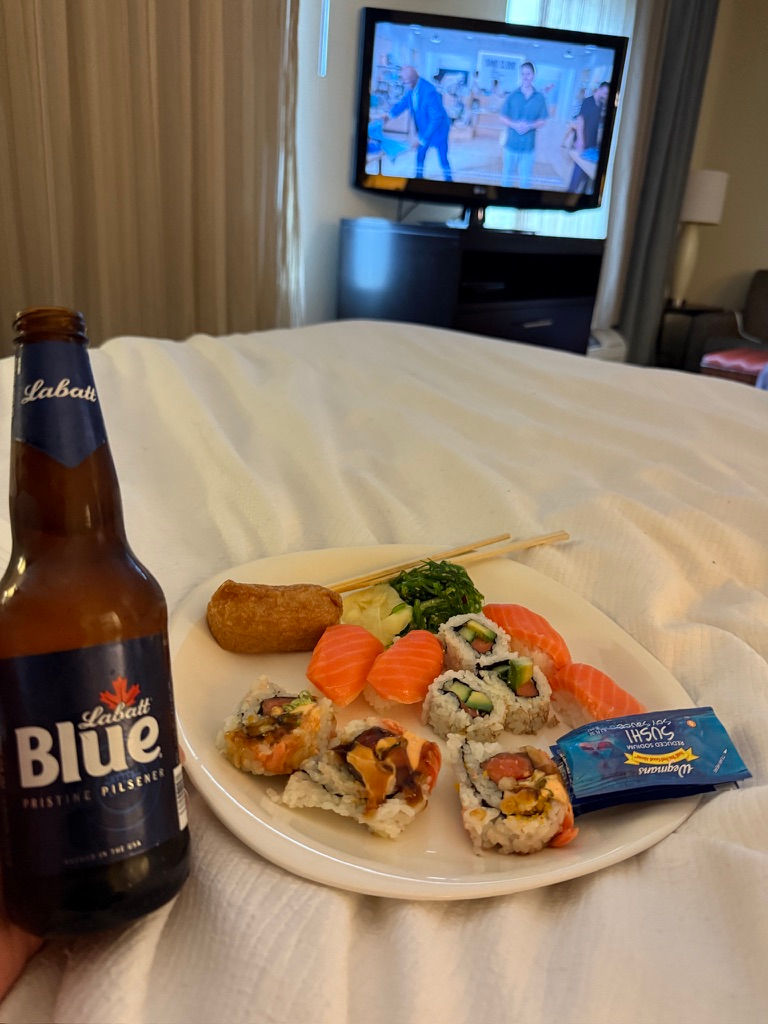 Beer and sushi on a bed with a TV showing a show in the background. Labatt Blue bottle, assorted sushi on a white plate, relaxed setting.