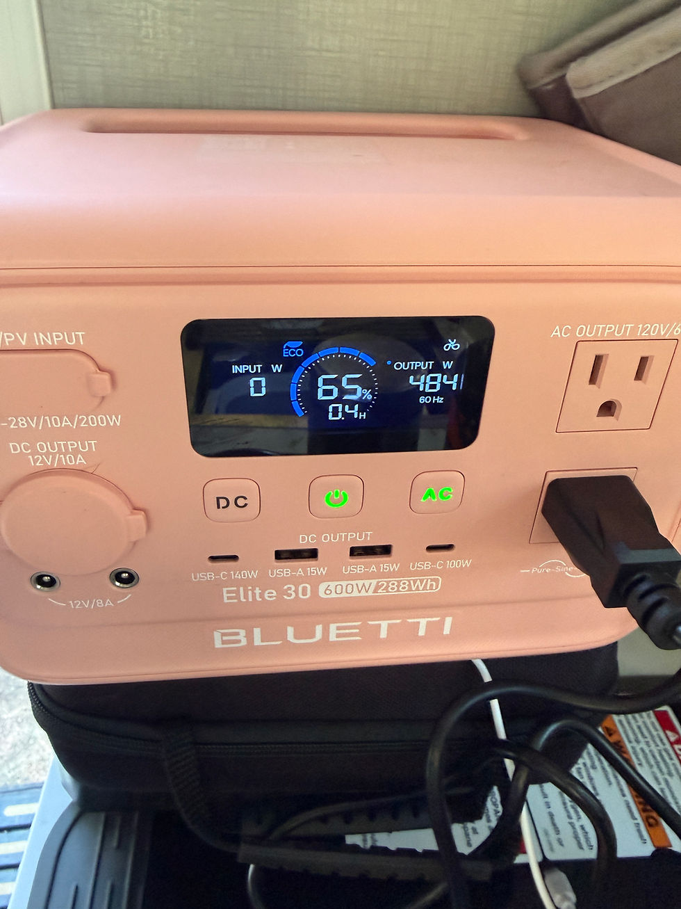 Pink Bluetti power station displaying 65% charge. Various input and output ports visible. Cords connected. Bright interior setting.