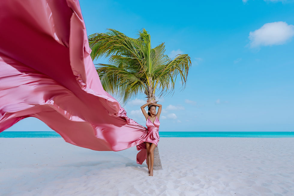 A woman on the beach leaning against a palm tree wearing a dusty rose pink flying dress