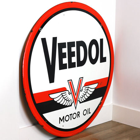 Large round Veedol Motor Oil porcelain sign with vibrant red outer border showing bold black VEEDOL lettering and winged V emblem leaning against white wall