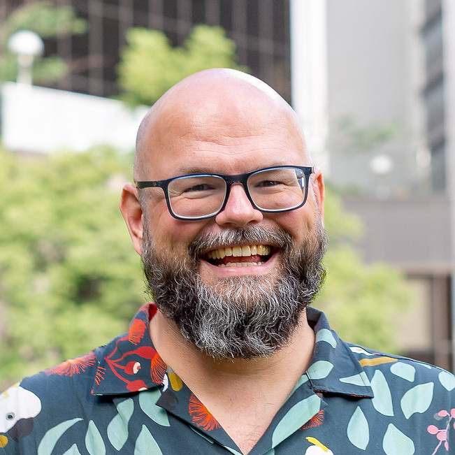 James. A man with no hair, glasses and a beard. He is smiling and wearing a shirt with cockatoos on it.