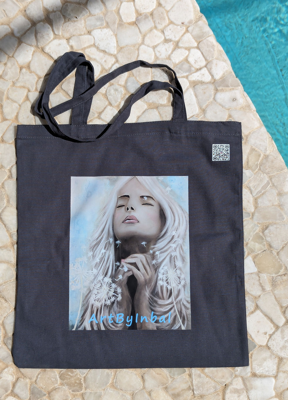 תיק בד קנבס בצבע שחור עם הדפס אומנותי של המותג "ArtByInbal". ההדפס מציג דמות של אישה עם שיער בלונדיני ארוך, עיניה עצומות, ופנ