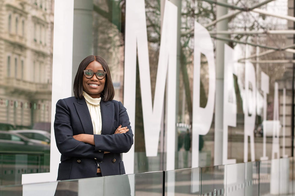 Smiling woman with glasses and arms crossed stands in front of glass wall with large "IMPERIAL" text. Urban setting with reflections.