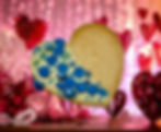 Gluten-free heart-shaped biscuit, half-dipped in Belgian white chocolate and decorated with vibrant blue sprinkles, displayed against a romantic pink and red backdrop with heart-shaped decorations and soft lighting