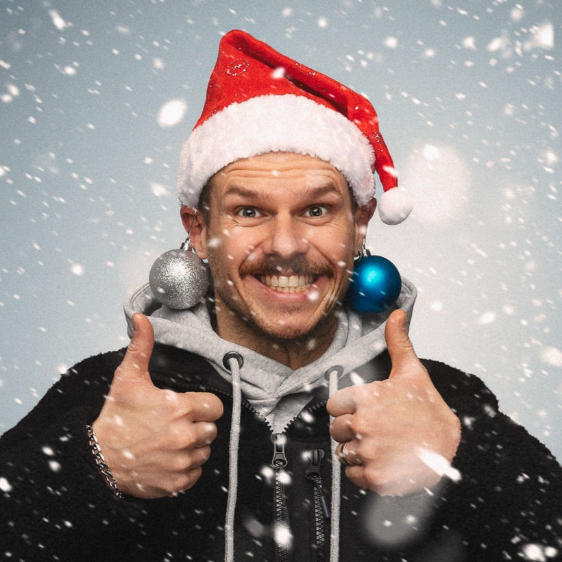 Lustiger Mann mit Oberlippenbart und Nikolausmütze beim Winter Shooting mit Schneefall. Er hat auch Christbaumkugeln an den Ohren hängen.