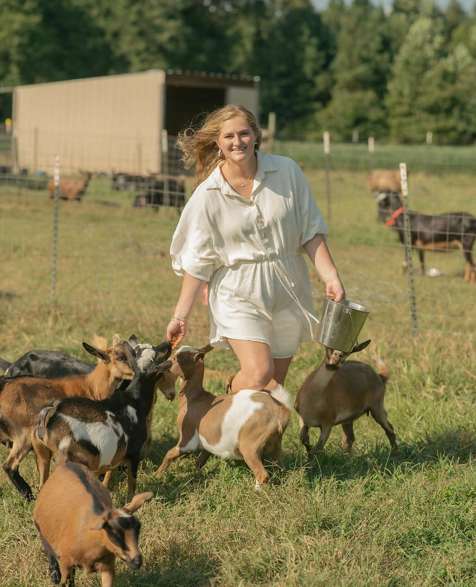 Smiling woman in white dress feeds goats from a metal pail in a sunny pasture near a barn.