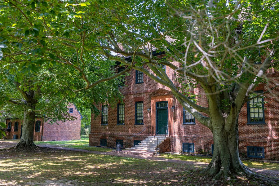 Historic brick building with green door set among lush trees. Sunlight filters through leaves, casting patterns on the grassy yard. Calm ambiance.
