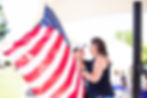 Two women embrace beside a large U.S. flag at an outdoor event under a white canopy, with a warm, celebratory mood.