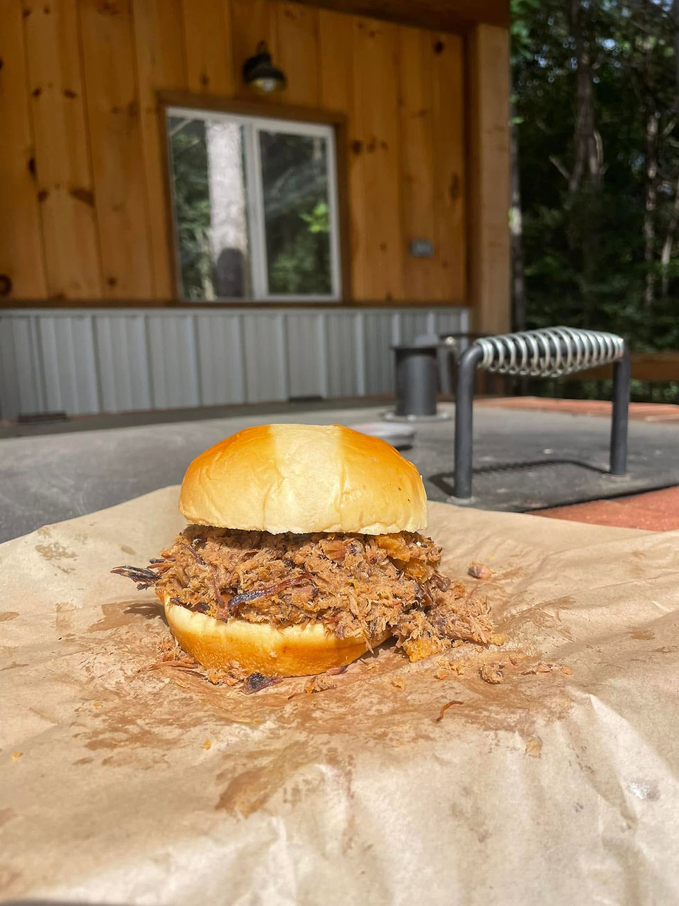 Pulled pork sandwich on a bun on wax paper, set on an outdoor porch with wood siding and trees in the background.