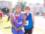 Two smiling women in race gear with medals stand at a sunny finish line, one in blue suit and one in pink visor and sunglasses