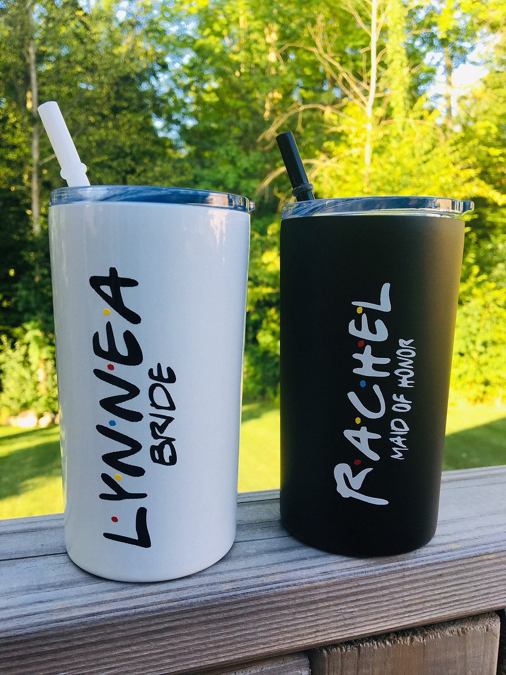 photo of two tumblers (one white one black) with wording similar to the "Friends" theme sitting on a wooden railing. Lettering is enlarged for detail and trees can be seen in the background. All Via Unmeasured Events