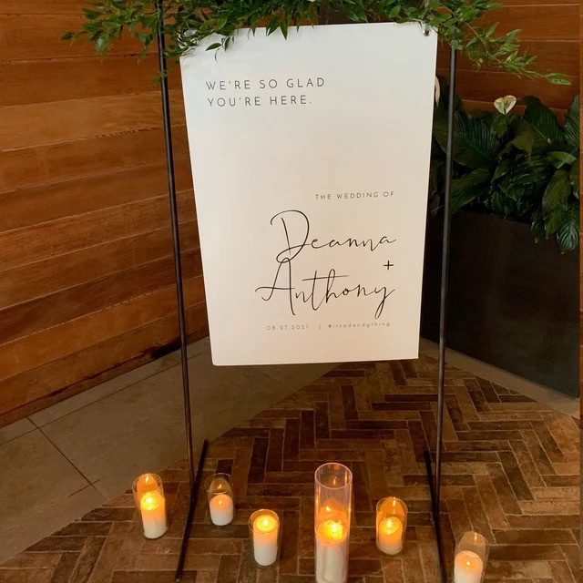 Photo of ADELLA custom wedding welcome sign that features a signature font and minimalist, clean lines. There is greenery surrounding the sign and candles can be seen at the front of the photo. Stationery from Unmeasured Events