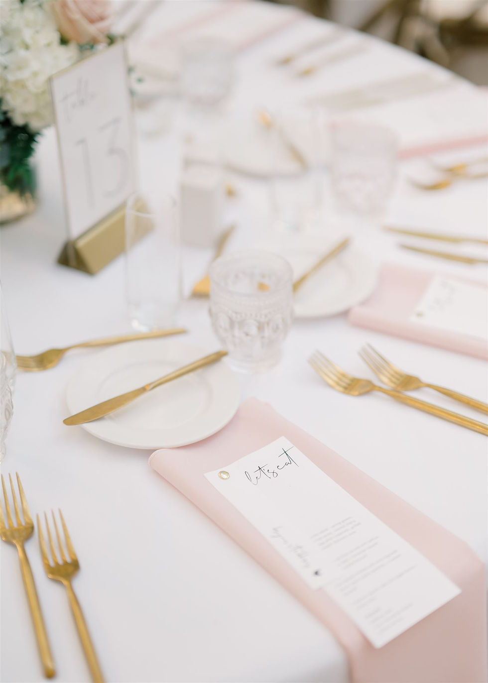 ADELLA "let's eat" menu on a pink napkin along with all white and gold settings on a wedding reception table. all via unmeasured events