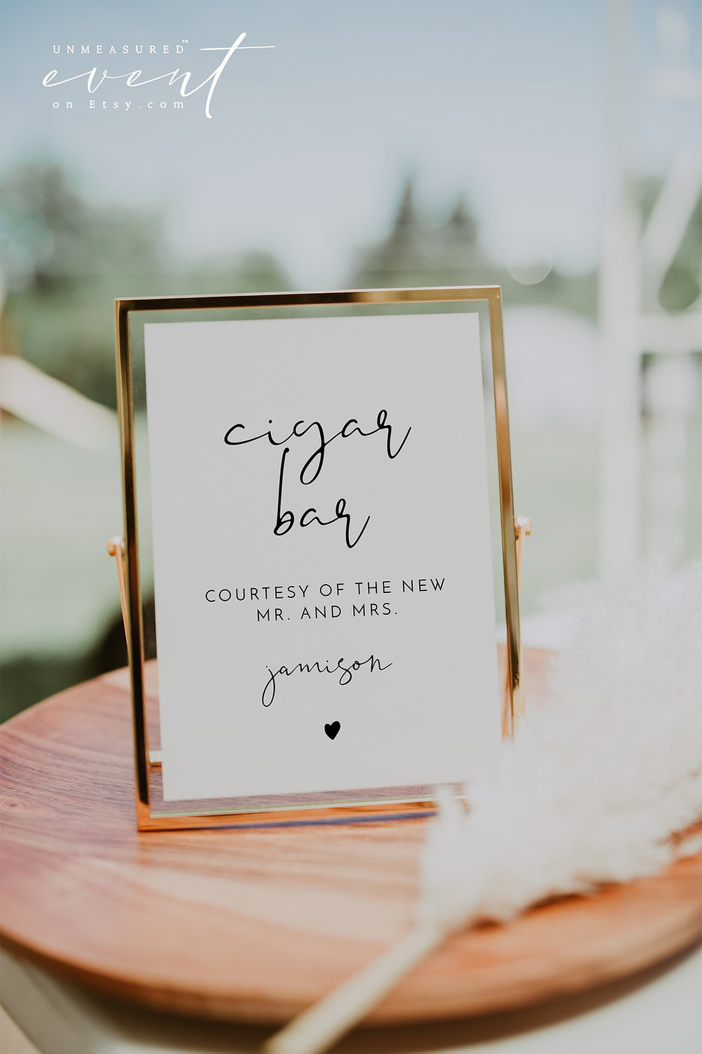 ADELLA Collection cigar bar sign written in an elegant script font that has a modern minimalist design, and a small little heart at the bottom. sign is in a gold frame and is sitting on a wooden table. All via unmeasured events