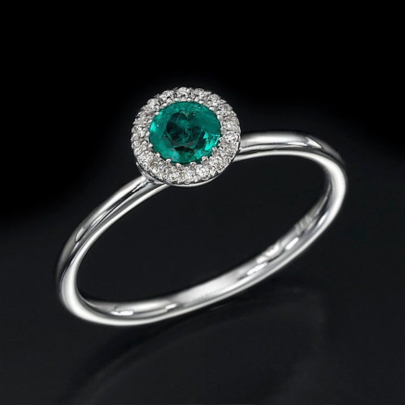 photo of a circular green emerald ring that has diamonds circling the emerald. The band is in white gold and the stone is tilted up to show detail