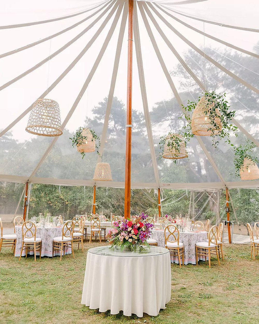 photo of a clear top tent and underneath wedding reception tables and various florals. A rainy sky and trees can be seen in the background, almost in a fog. All via unmeasured events