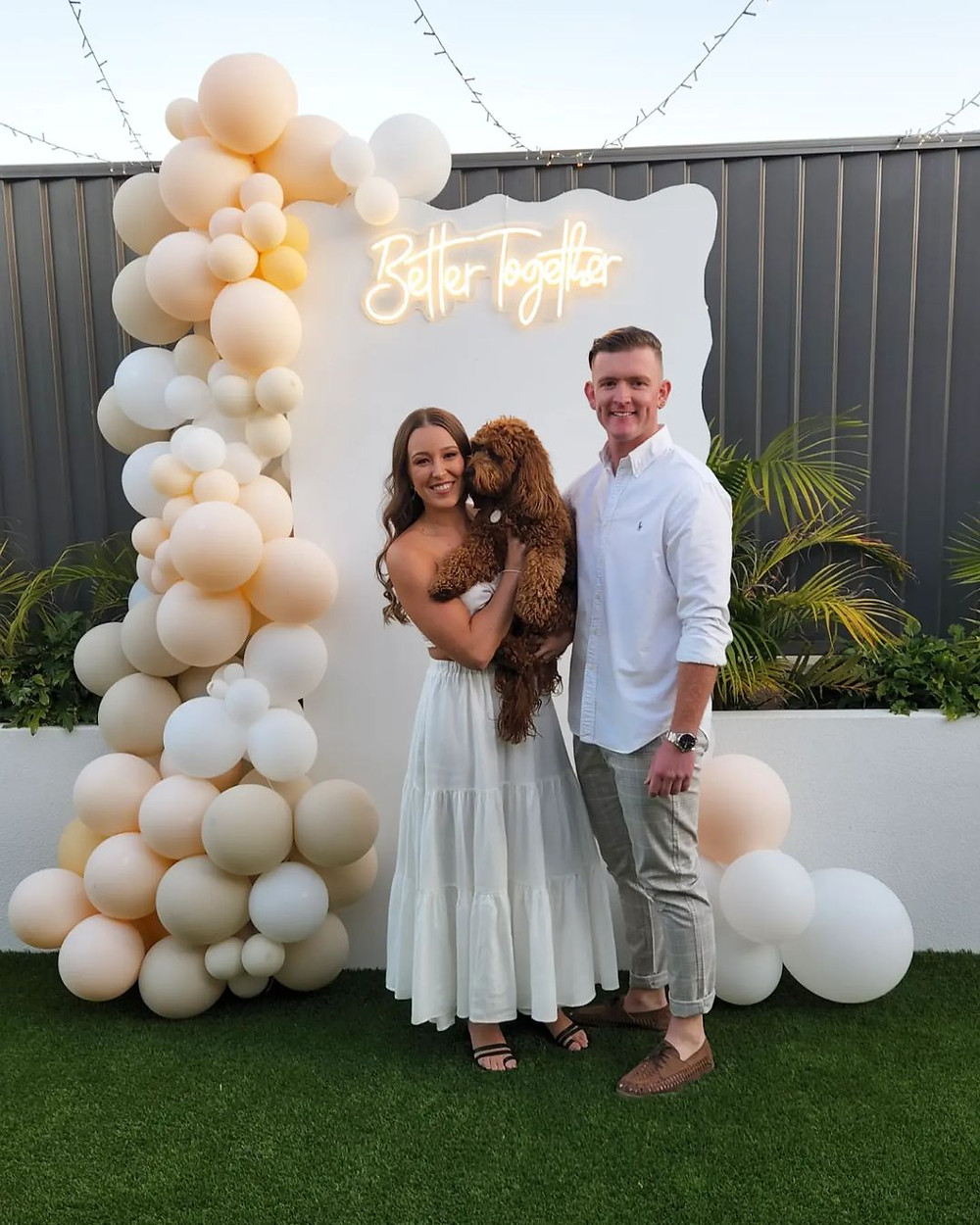 photo of a newly engaged couple standing in-front of a white "better together" wall at their engagement party as they wear all white and bride holds their dog. all via unmeasured events.